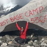 Female trekker posing at Sagarmatha (Everest) Base Camp location with prayer flags, snowy peaks, and the Khumbu Glacier during the Everest Base Camp trek in Nepal
