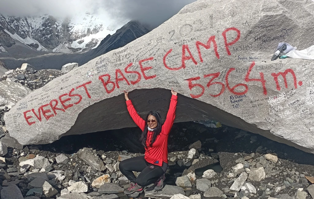 Female trekker posing at Sagarmatha (Everest) Base Camp location with prayer flags, snowy peaks, and the Khumbu Glacier during the Everest Base Camp trek in Nepal
