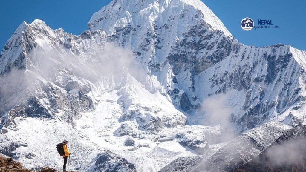 A trekker stands on a rocky mountain ridge, wearing hiking gear and a backpack, looking out at a vast Himalayan panorama. The main subject is in the foreground on the left, while the iconic, snow-capped pyramid of Mount Everest is clearly visible in the distance against a clear blue sky, identifying where Mount Everest located.