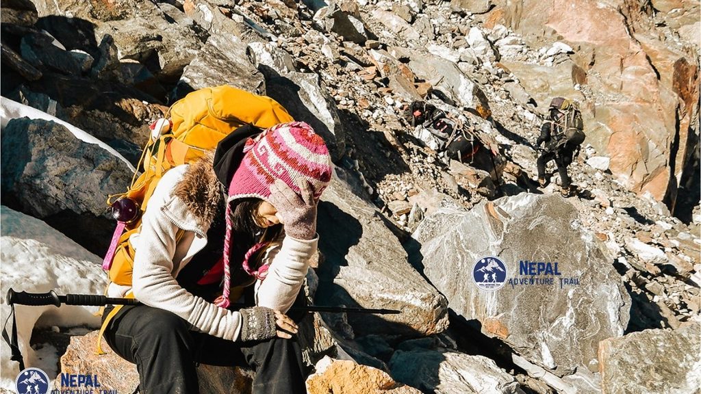 A female trekker in warm outdoor clothing sitting on a rocky trail with her hands holding her head in pain, illustrating the symptoms associated with Everest Base Camp altitude sickness.