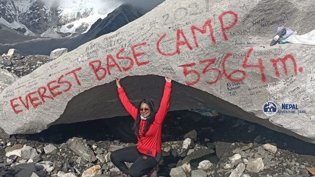 A female trekker in a red and blue jacket and beanie smiling proudly next to the iconic Everest Base Camp stone monument, which is draped in colorful prayer flags with the Khumbu Glacier and snow-capped Himalayas in the background.