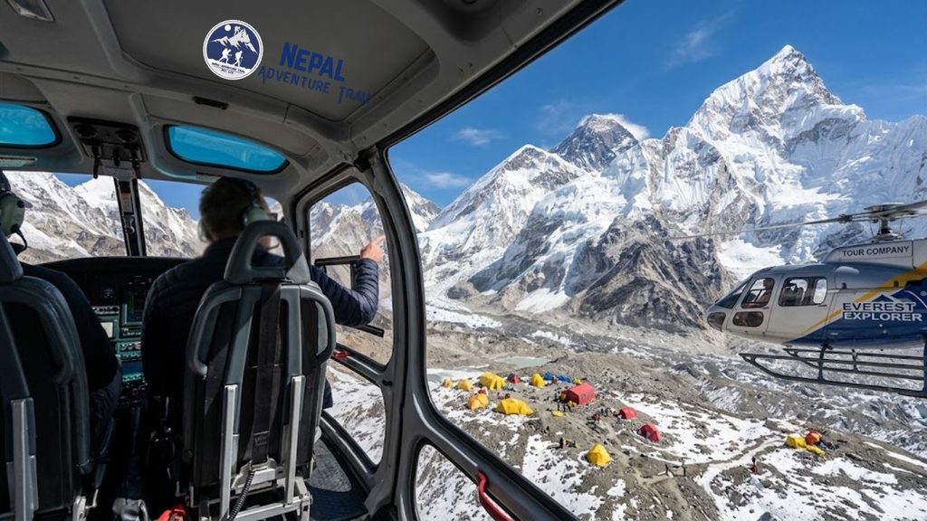 Panoramic view from inside a helicopter looking out the window at Mount Everest, the Khumbu Glacier with yellow Base Camp tents, and a second helicopter flying in the high Himalayas during an Everest Base Camp Helicopter Tour.