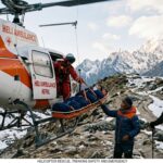 A high-altitude rescue operation by Heli Ambulance Nepal in the Himalayas. A rescue worker in a red flight suit, suspended by a hoist, lowers an injured trekker in a orange basket stretcher to the ground. A local Nepali guide in a blue jacket stabilizes the stretcher. A Western trekker with a large backpack and poles watches nearby. Snow-capped mountains, rugged terrain, and prayer flags fill the background.