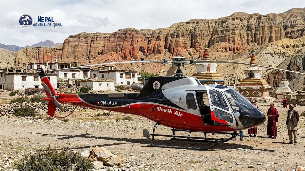 Travelers stand near a parked red and blue commercial helicopter labeled 'MUSTANG HELI' and 'FLY NEPAL' on a remote mountain helipad in Upper Mustang, with snow-capped Himalayan peaks, arid canyons, and a traditional village in the background during a Mustang Helicopter Tour Nepal.