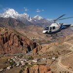 Aerial view from a Mustang Helicopter Tour Nepal flying over the arid, red rock canyons and traditional villages of Upper Mustang with the snow-capped Himalayan peaks in the background.